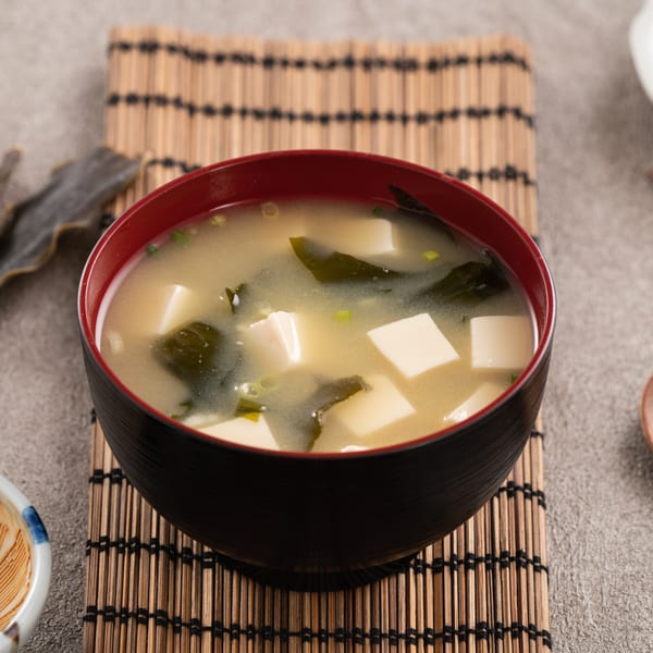 A black and red bowl of miso soup with tofu and seaweed on a bamboo mat.