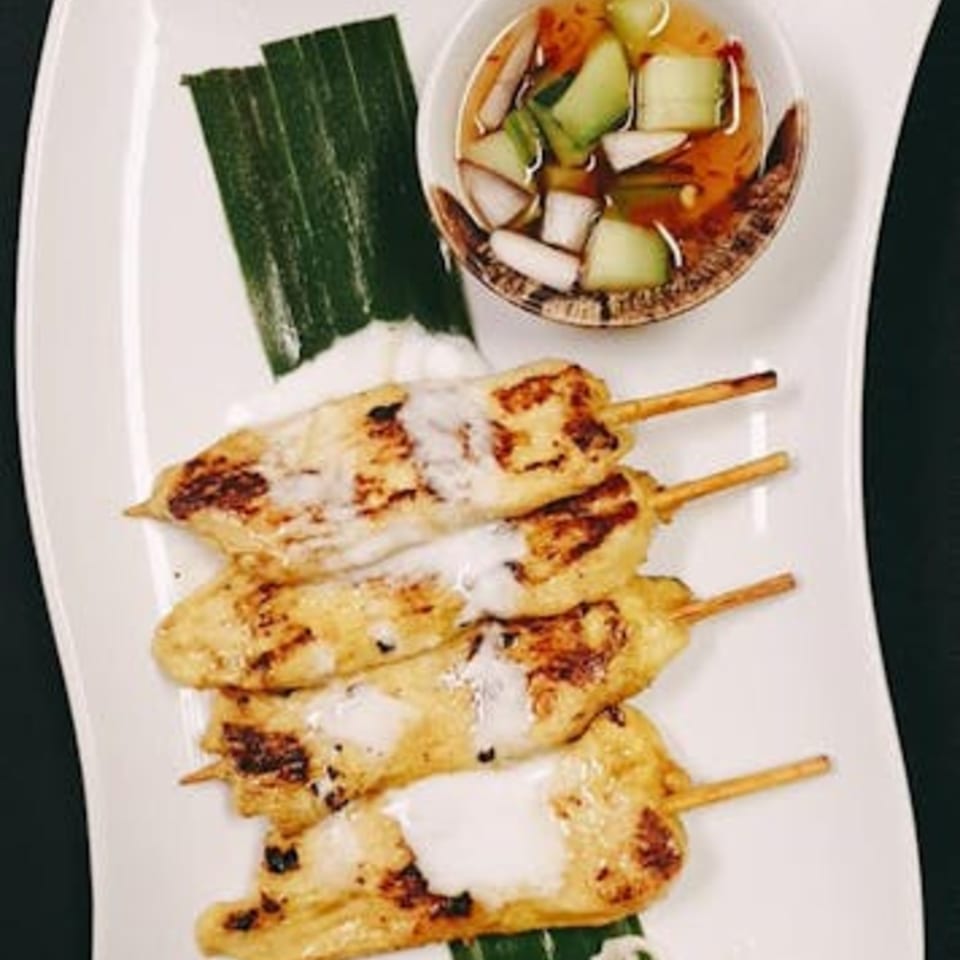 Grilled chicken satay skewers with coconut cream drizzle, served with cucumber relish on a white plate.